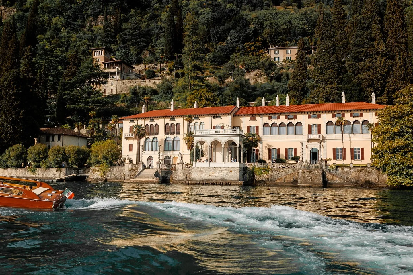 Villa with red-tiled roof and arched windows on a lakeshore surrounded by dense greenery, with a wooden boat passing in the water.