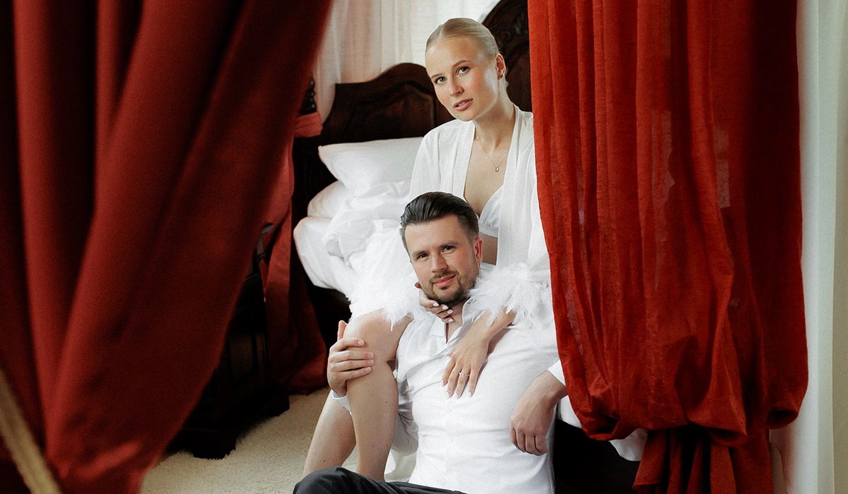 Man and woman in white robes sitting closely on a bed surrounded by red curtains.