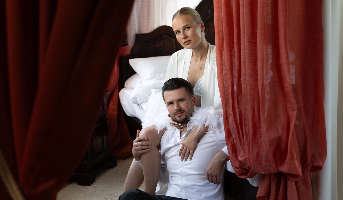 Couple in white attire sitting by a dark wooden bed with red curtains in the foreground.