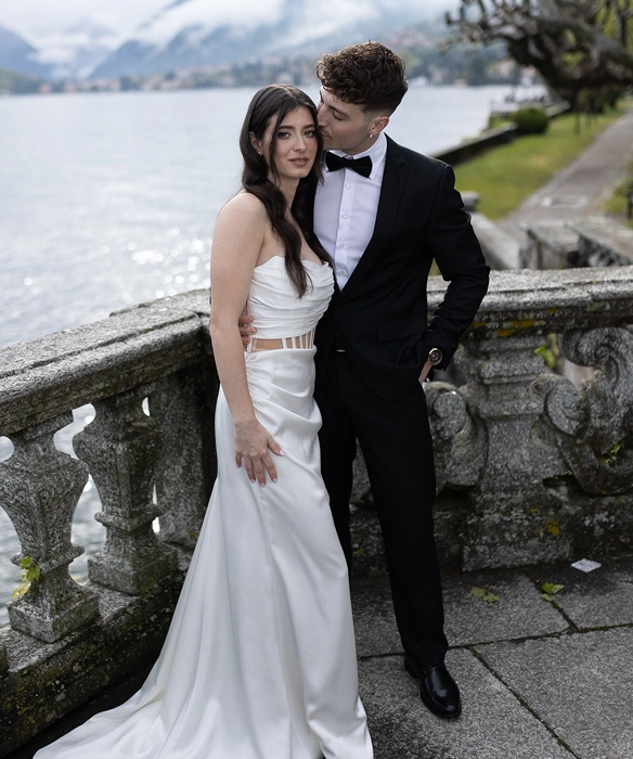Couple dressed formally by a stone railing with a lake and mountains in the background, the man kissing the woman's temple.