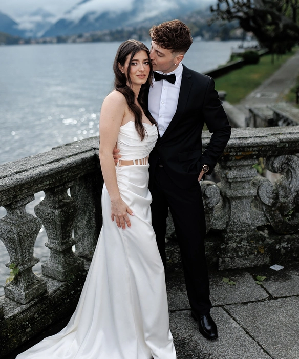 Couple dressed formally with the woman in a white dress and the man in a black tuxedo, standing by a stone railing by a lake.