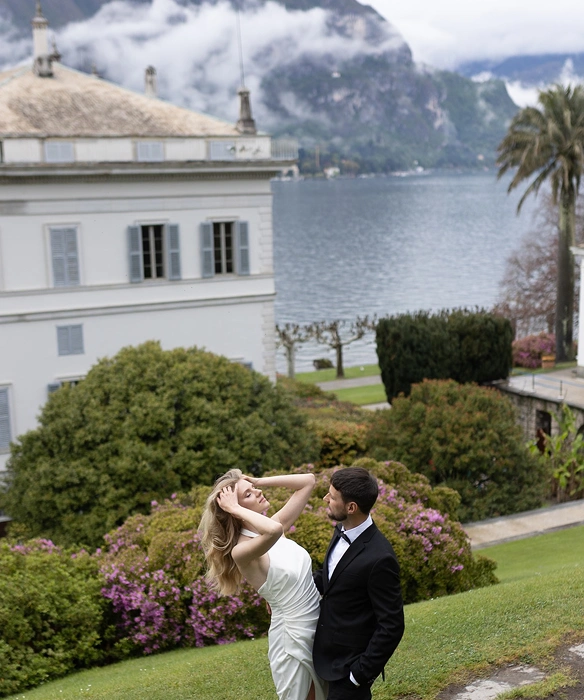 Couple dressed formally with the woman in a white dress and the man in a black tuxedo standing on green grass near flowering bushes, with a large building and a lake with mountains in the background.