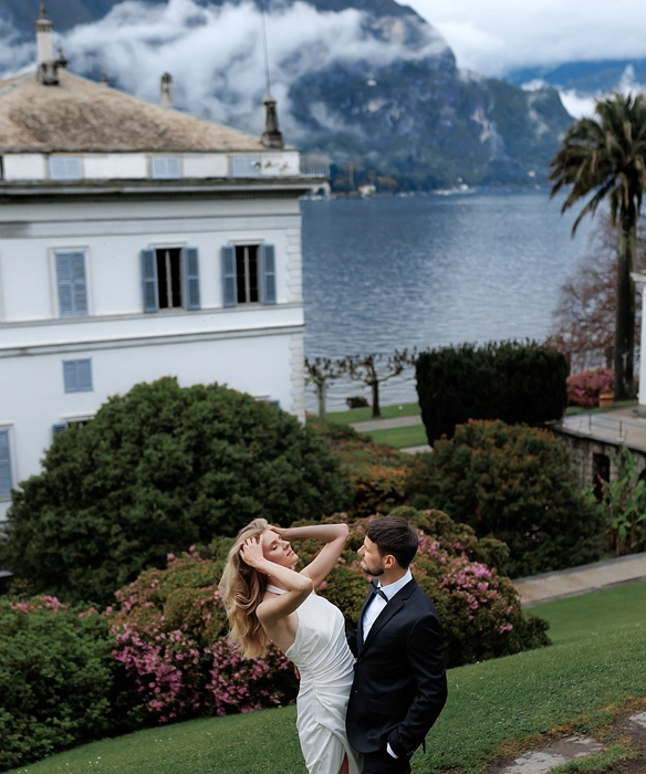 Woman in white dress and man in black suit posing on a green lawn with flowering bushes, a white building, lake, and misty mountains in the background.