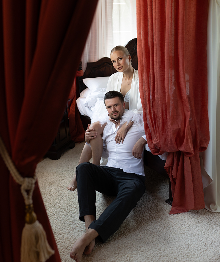 A couple in white attire seated on the floor by a bed with red curtains partially framing the scene.