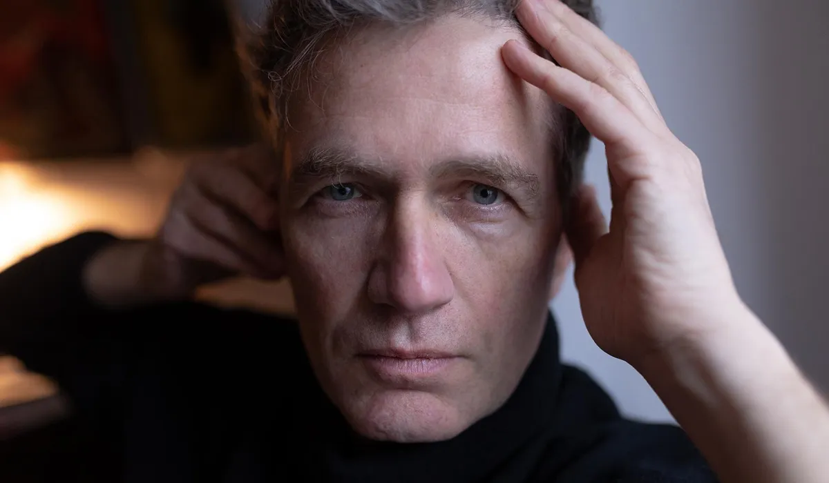 Close-up of a middle-aged man with blue eyes and light skin wearing a black shirt, touching his forehead with one hand.