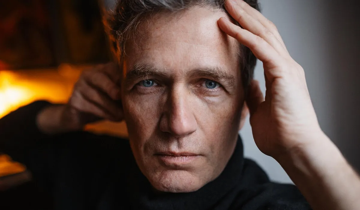 Close-up of a serious middle-aged man with blue eyes, holding his forehead with one hand and touching his neck with the other.