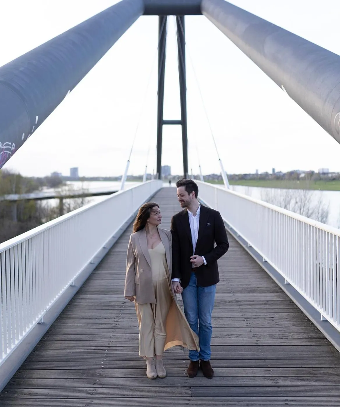 Couple holding hands and walking on a modern suspension bridge with river and cityscape in the background.