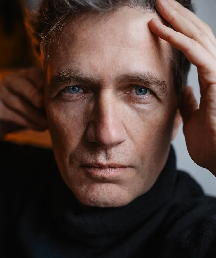 Close-up of a man with blue eyes and light skin wearing a black turtleneck, touching his head with both hands.