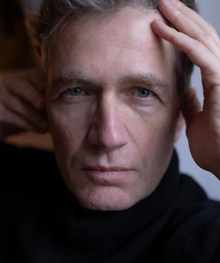 Close-up of a middle-aged man with light blue eyes and gray hair, wearing a black turtleneck, touching his forehead and ear.