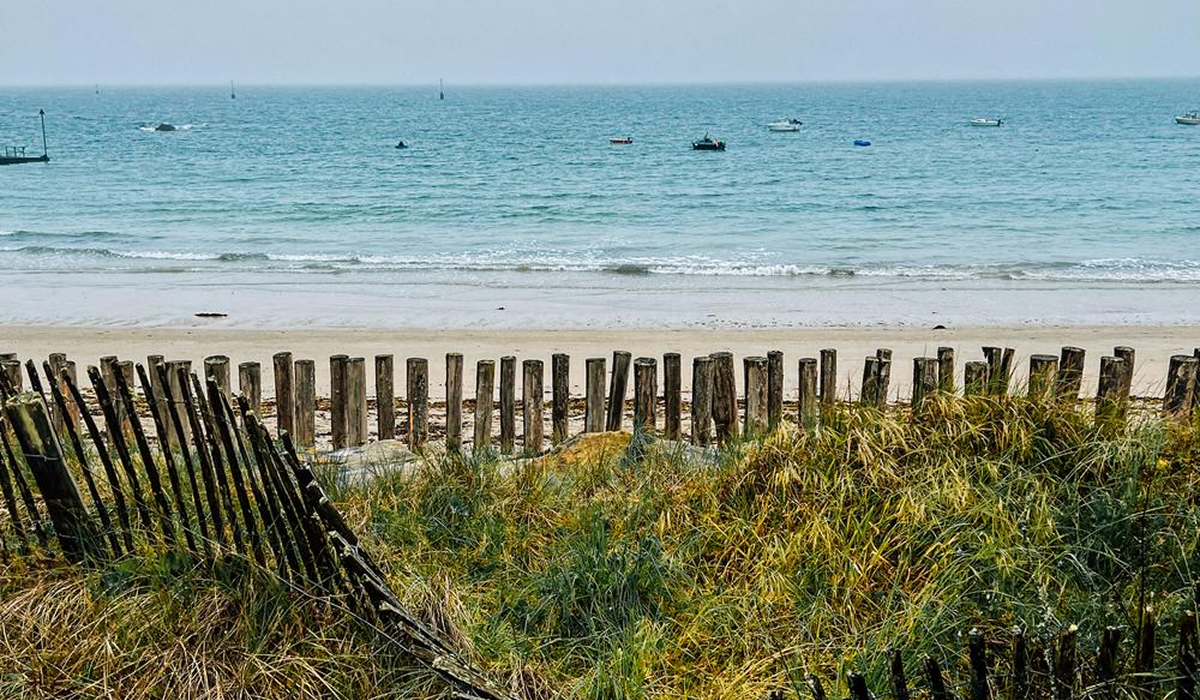 Calm ocean with small boats in the distance, sandy beach, wooden posts, and green beach grass in foreground.