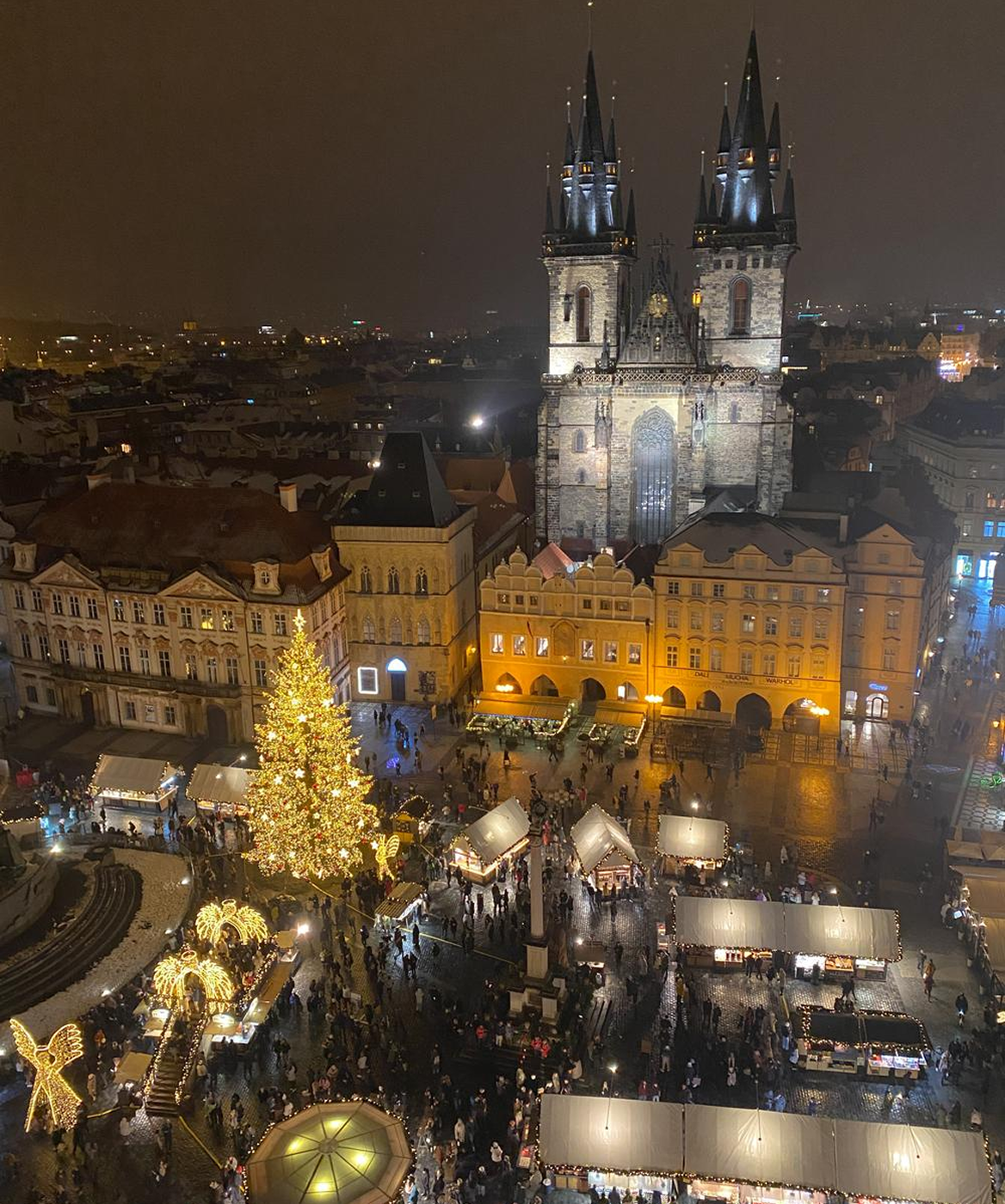 Aerial night view of a historic town square with a large illuminated Christmas tree, festive market stalls, and a Gothic church with tall spires in the background.