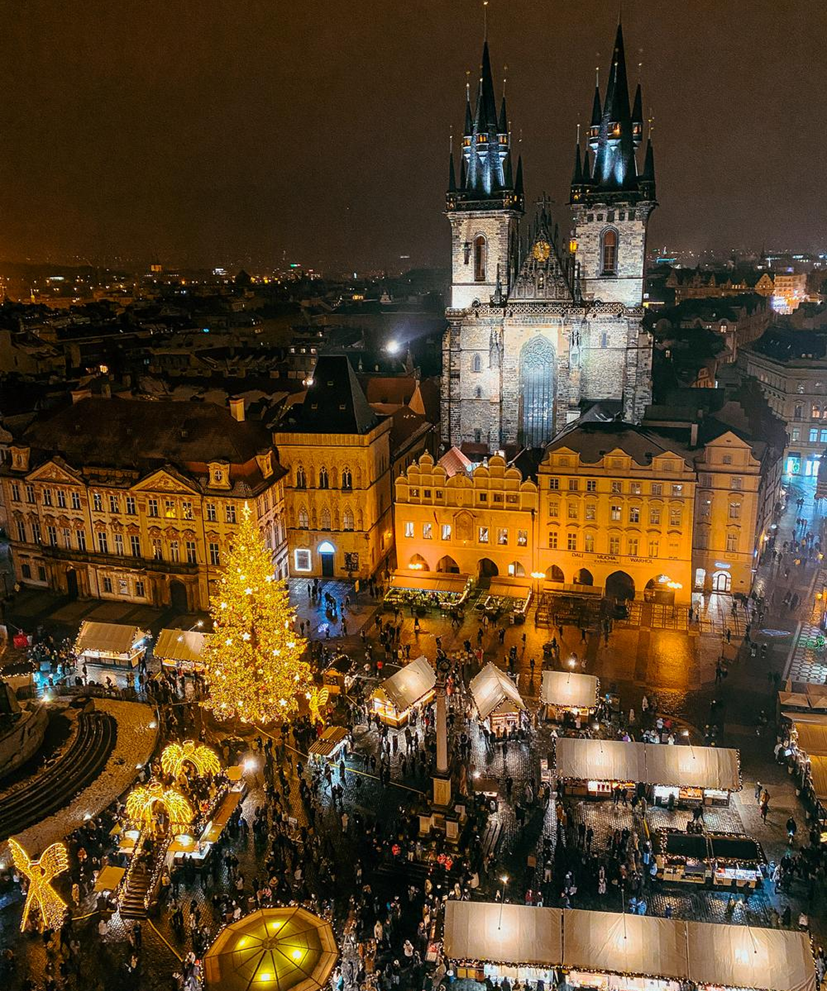 Night aerial view of a festive Christmas market with a large decorated tree and illuminated stalls in a historic European town square featuring a Gothic church.