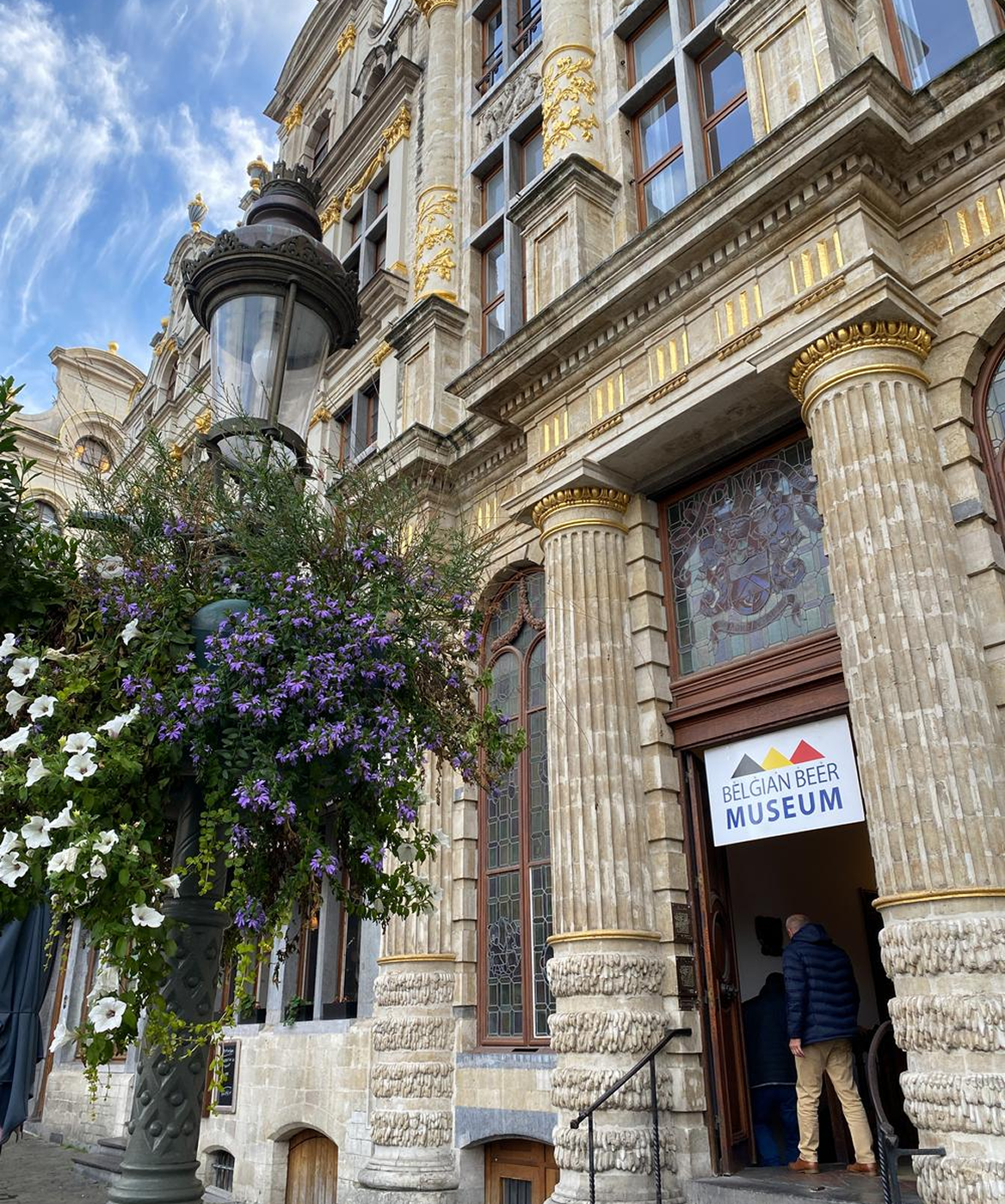 Entrance of the Belgian Beer Museum in a historic building with ornate columns and stained glass windows, a streetlamp with hanging flowers in the foreground.