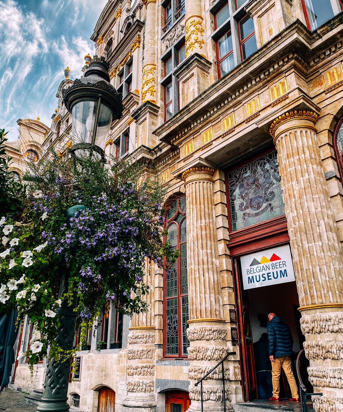 Entrance of the Belgian Beer Museum with ornate columns, stained glass windows, and a large hanging flower basket.