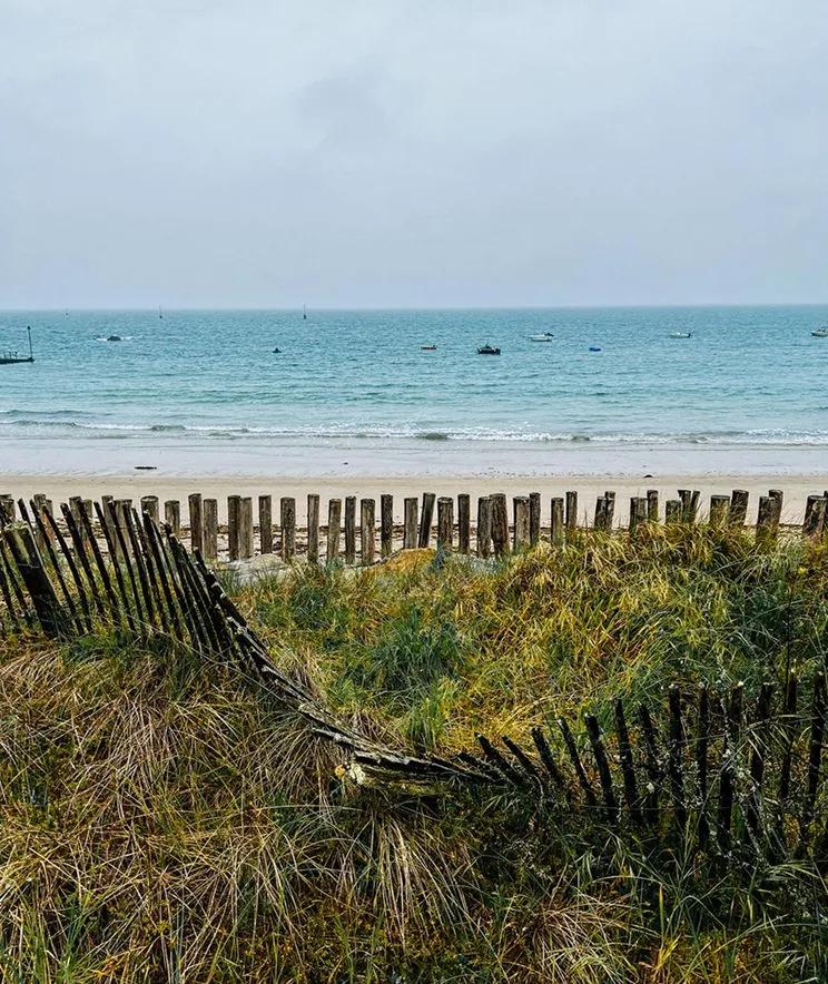 Ocean view with blue water and small boats in the distance, sandy beach, grassy dunes, and a weathered wooden fence in the foreground.