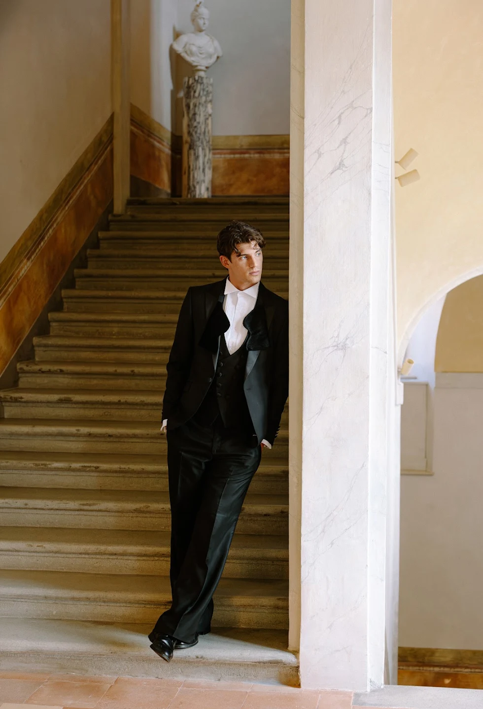 Man in black suit leaning against a white stone pillar with stairs and a marble bust in the background.