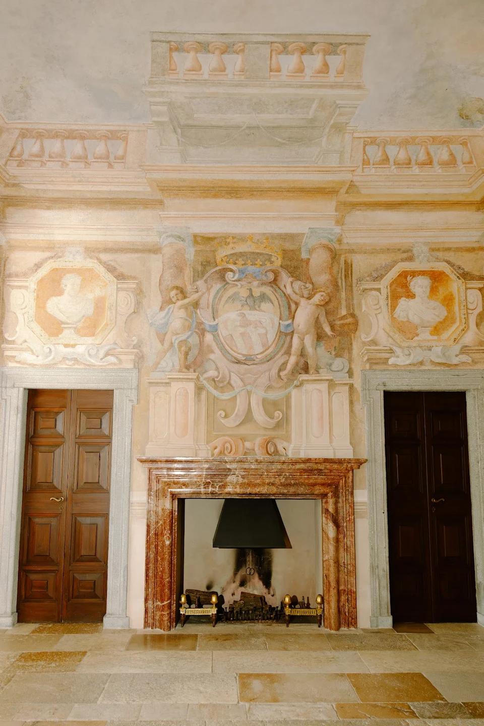 Ornate room wall with a marble fireplace flanked by two wooden doors and decorated with classical cherub frescoes and bust reliefs.