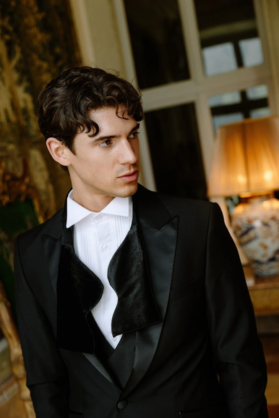 Young man with dark curly hair wearing a black tuxedo with a white dress shirt, standing indoors near a lamp and window.