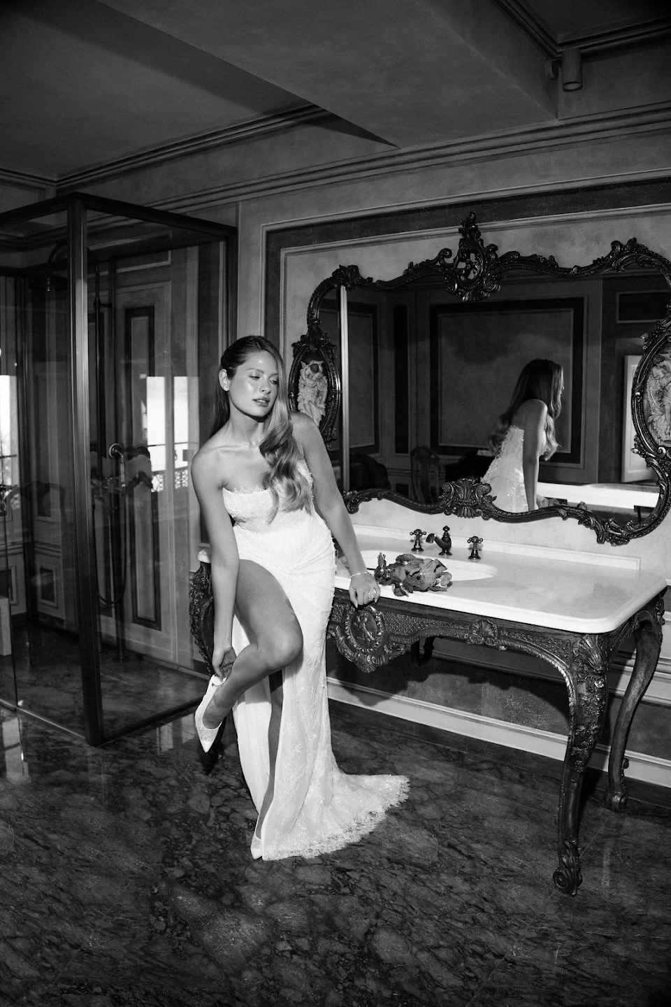 Woman in a white strapless gown with a high slit putting on a shoe while leaning on an ornate vanity with a large decorative mirror.