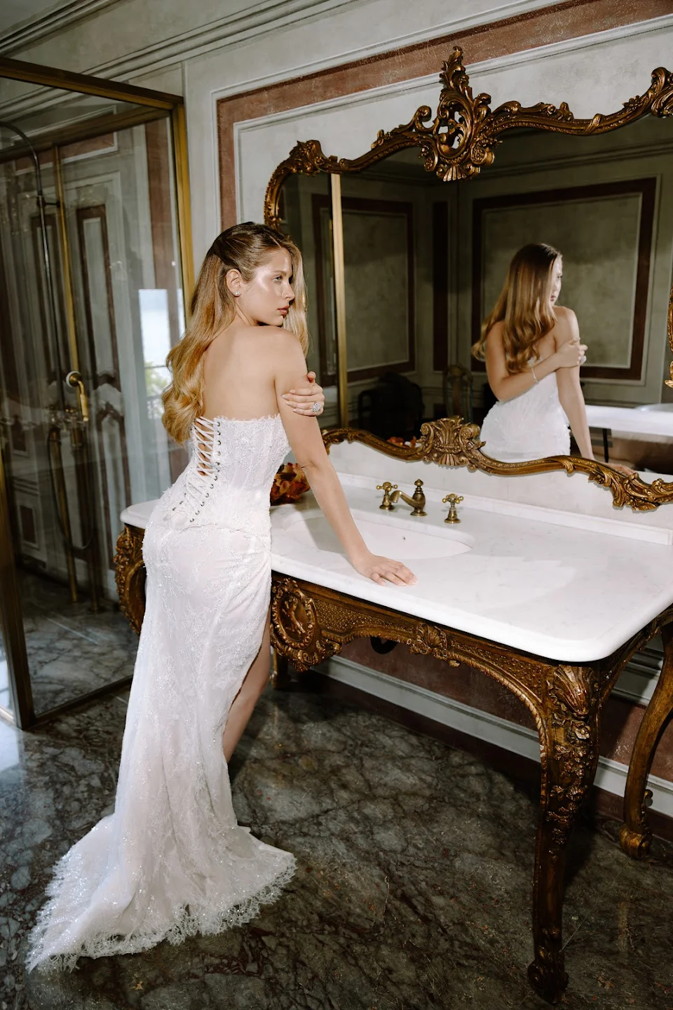 Woman in a white lace strapless gown standing in front of an ornate gold-framed mirror over a marble sink.