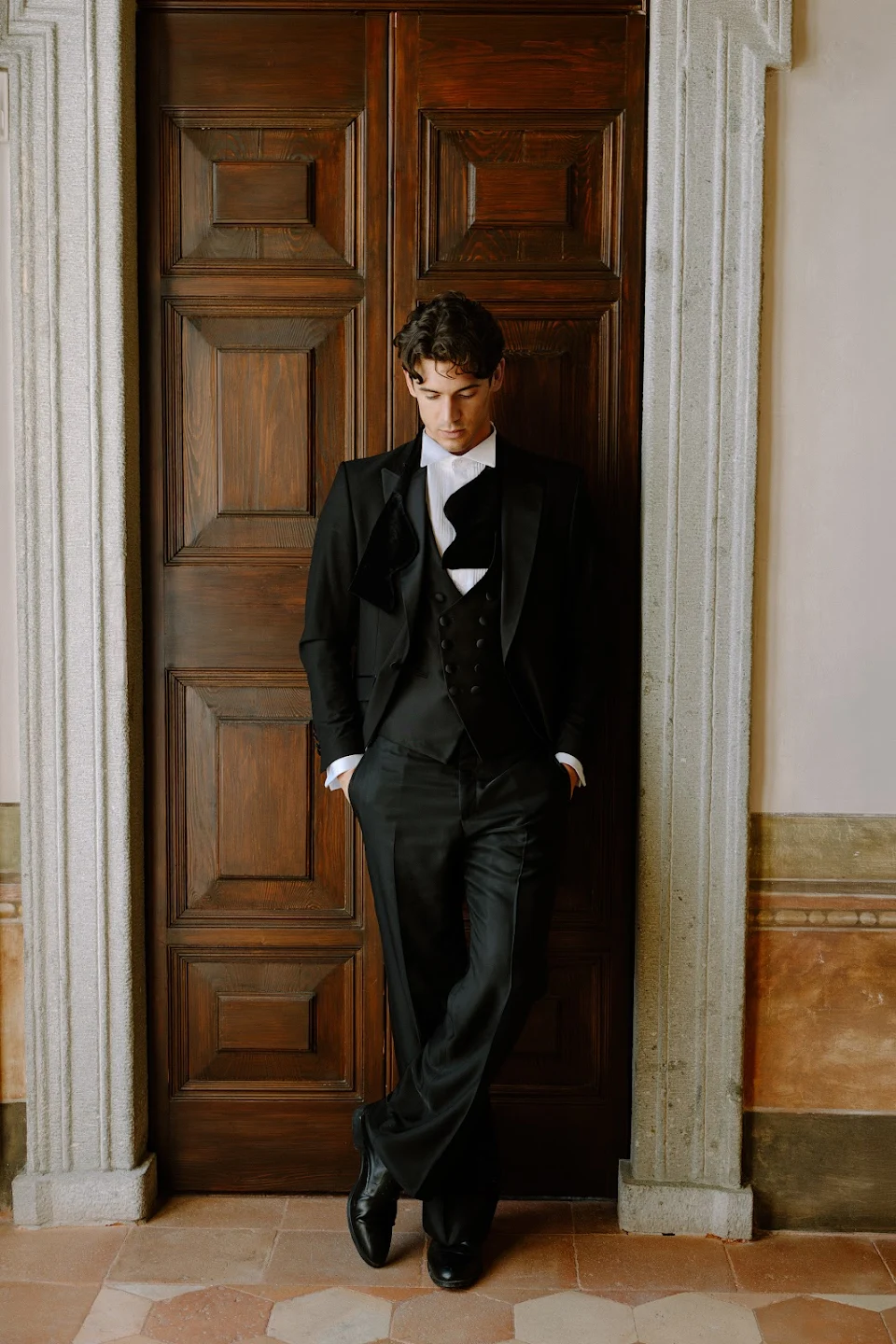 Man in a black tuxedo with a white shirt and black bow tie standing with hands in pockets, leaning against a large wooden door with stone frame.