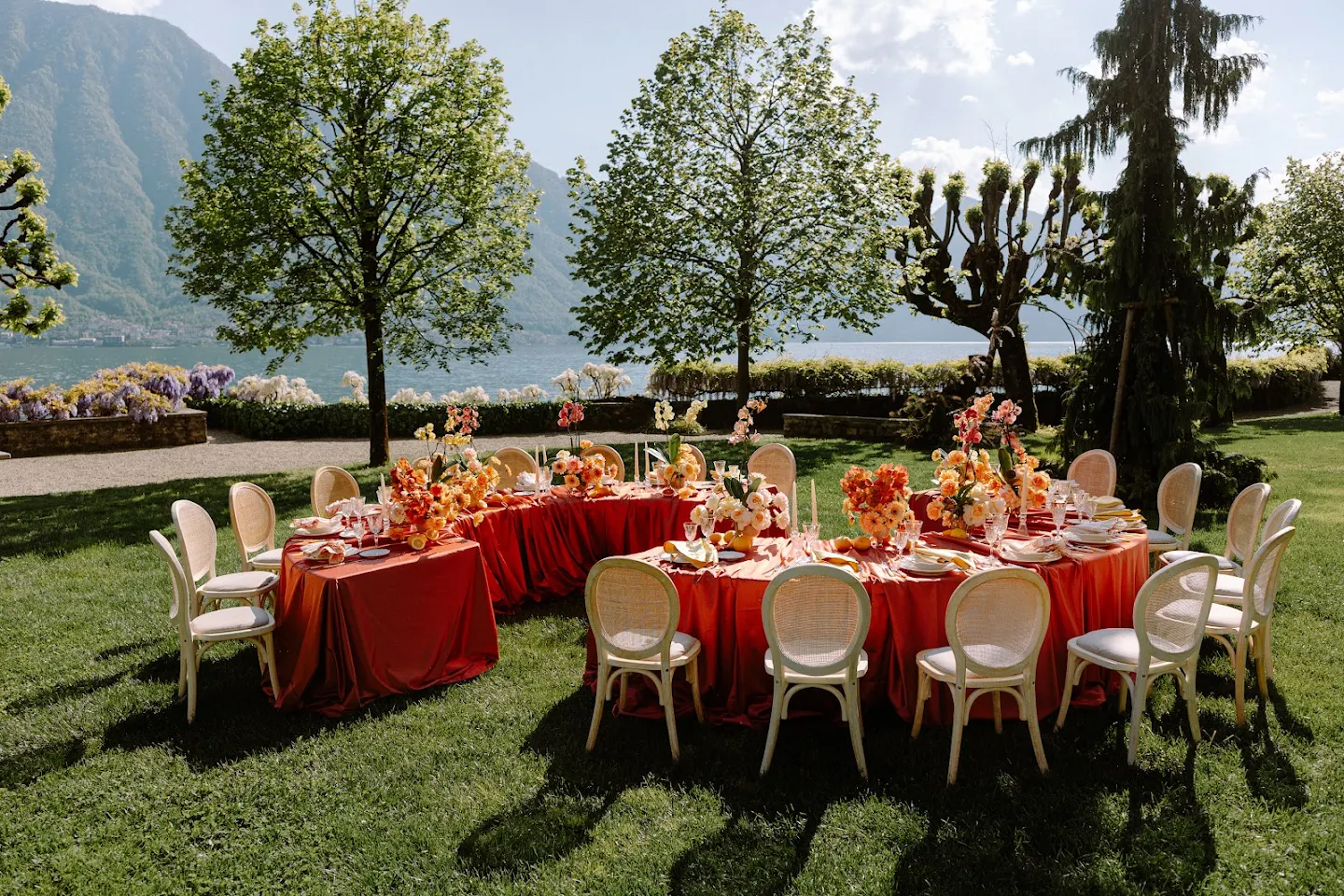 Outdoor U-shaped dining setup with red tablecloths, beige chairs, and orange floral centerpieces on green lawn by a lake with mountains in the background.
