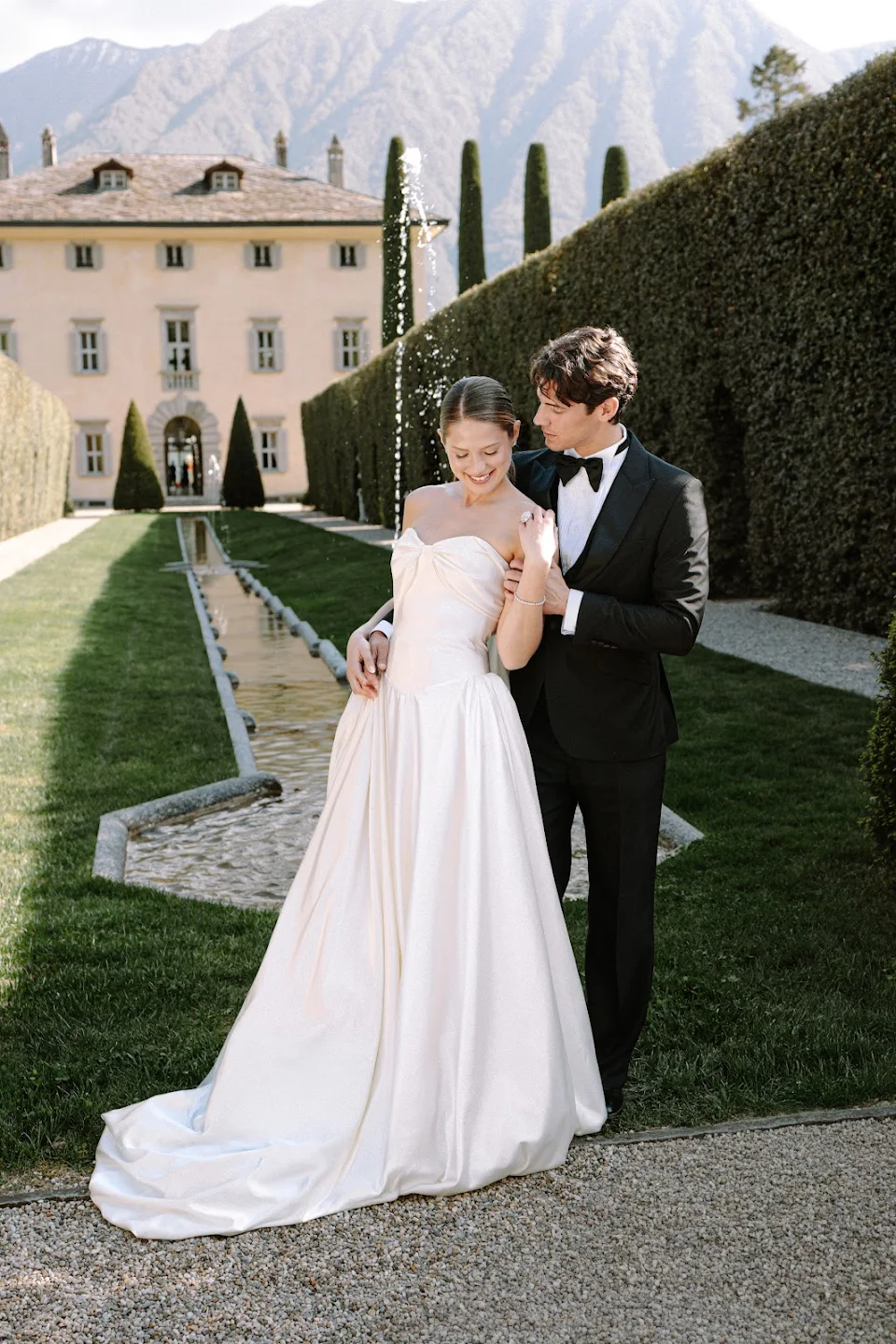 Bride in a white strapless gown and groom in a black tuxedo standing in a garden with a fountain and tall hedges.