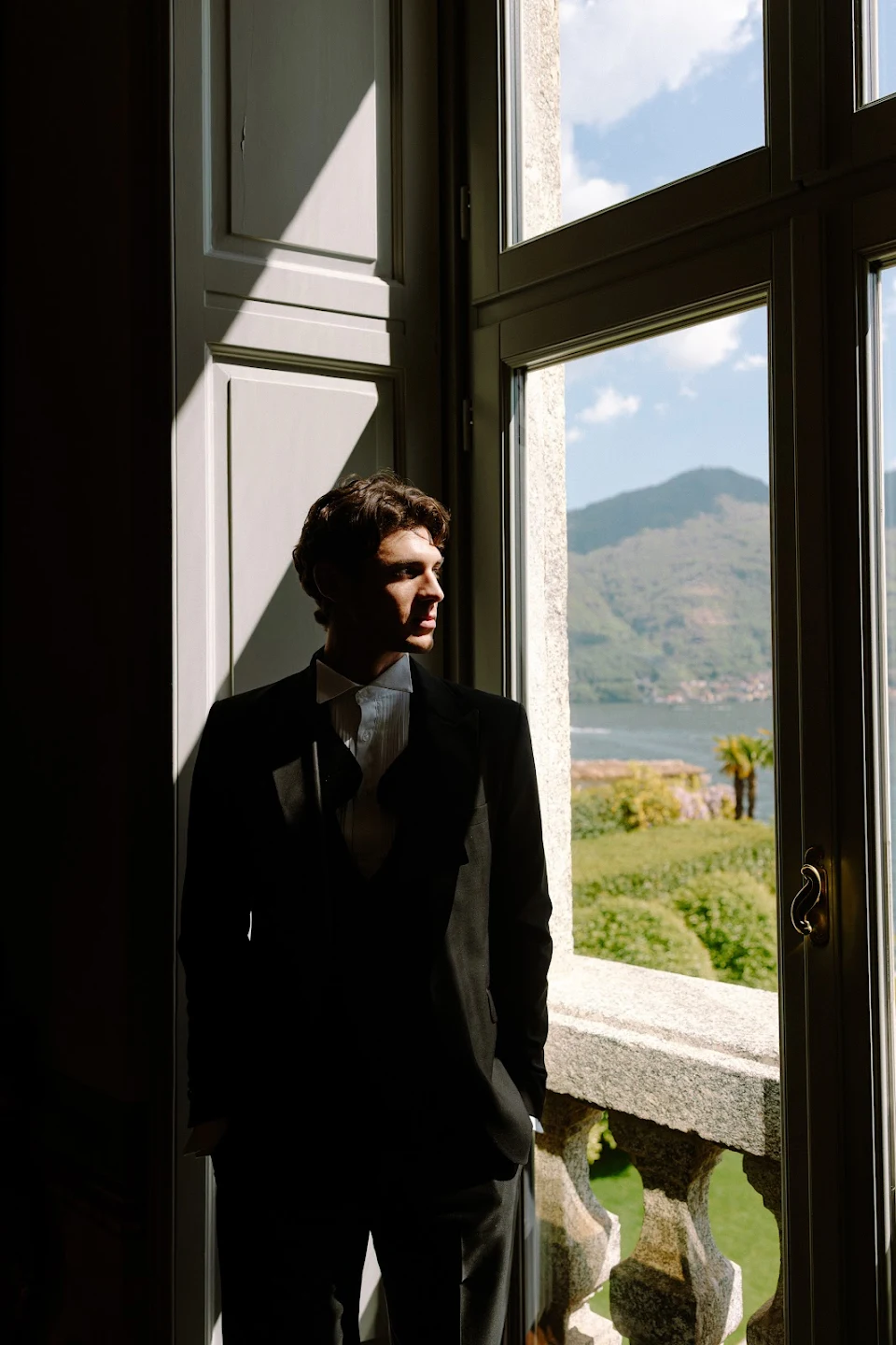 Young man in a black suit standing by a window with a scenic view of a garden and distant mountains.