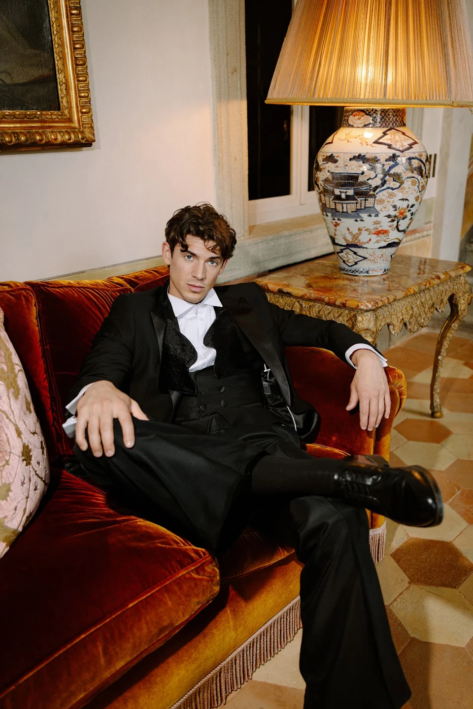 Young man in a black tuxedo lounging on a red velvet sofa in an ornate room with an antique lamp on a marble-top table.