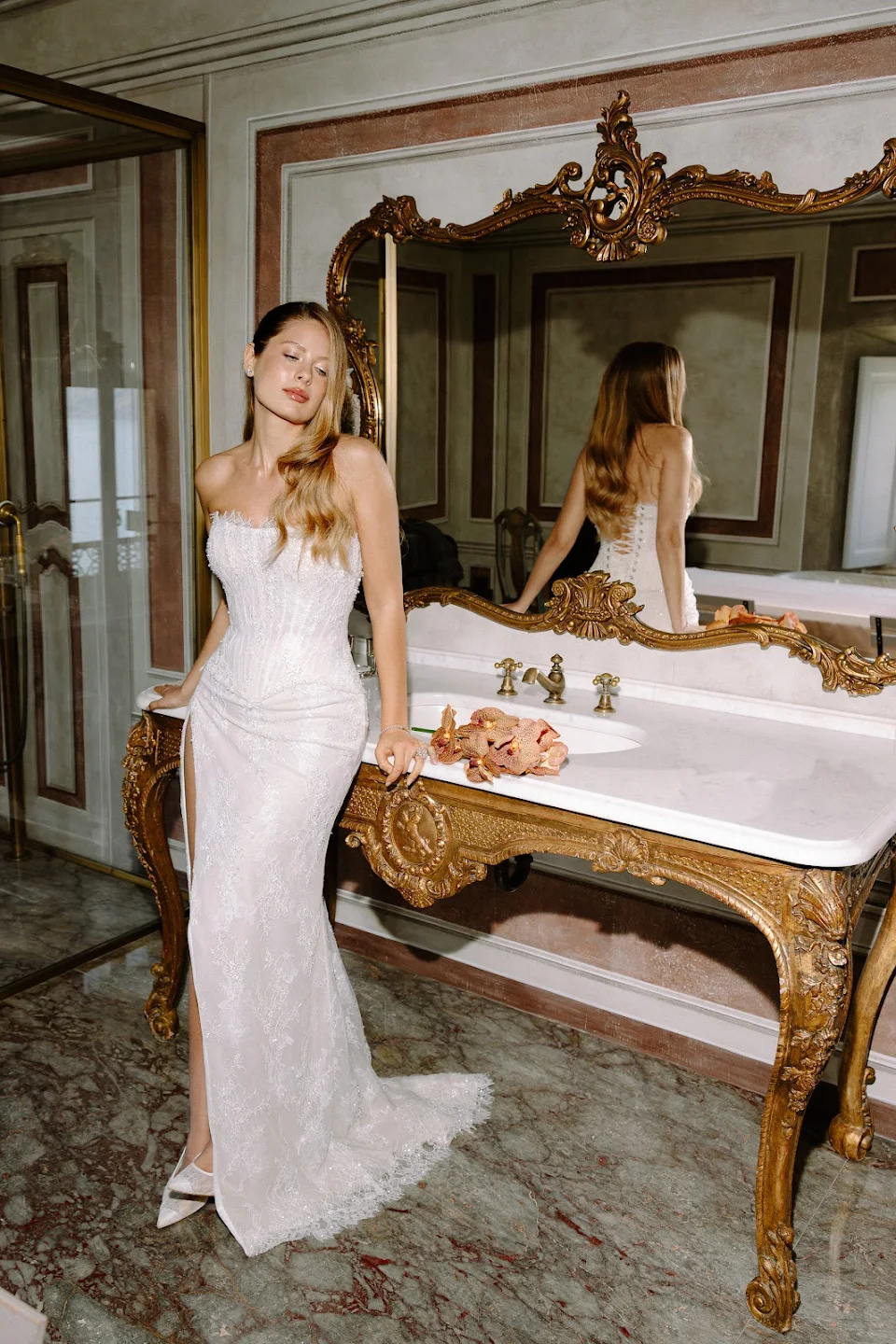 Woman in a white lace strapless gown with long wavy hair standing next to an ornate gold-framed mirror and antique sink in a marble-floored room.