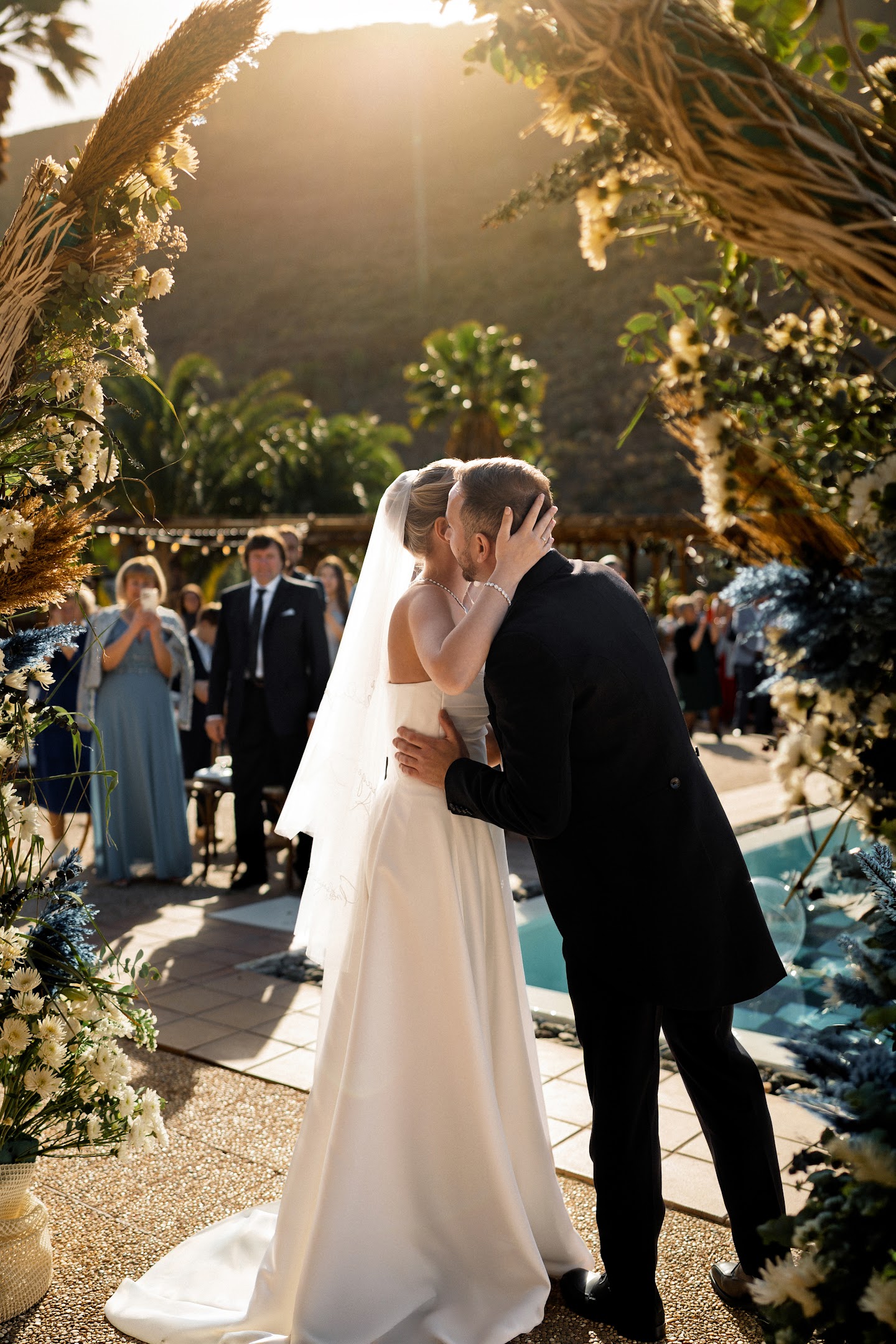 Bride and groom sharing a kiss during an outdoor wedding ceremony with guests in the background.