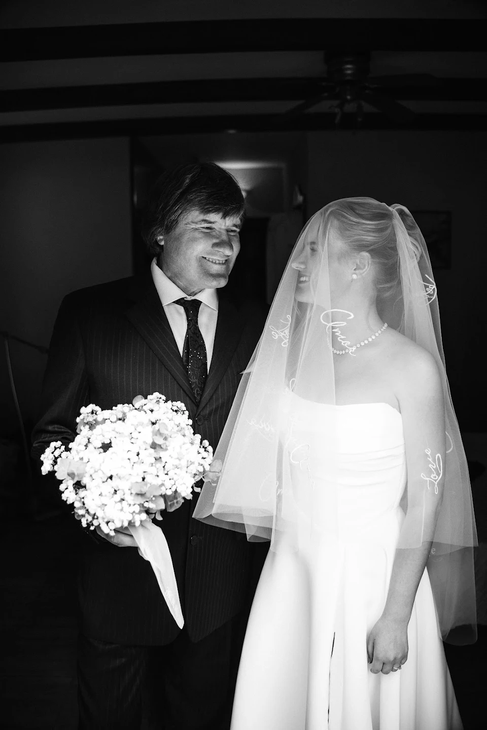 Black and white photo of a smiling bride in a wedding dress and veil holding the arm of a man in a suit who is holding a bouquet of flowers.