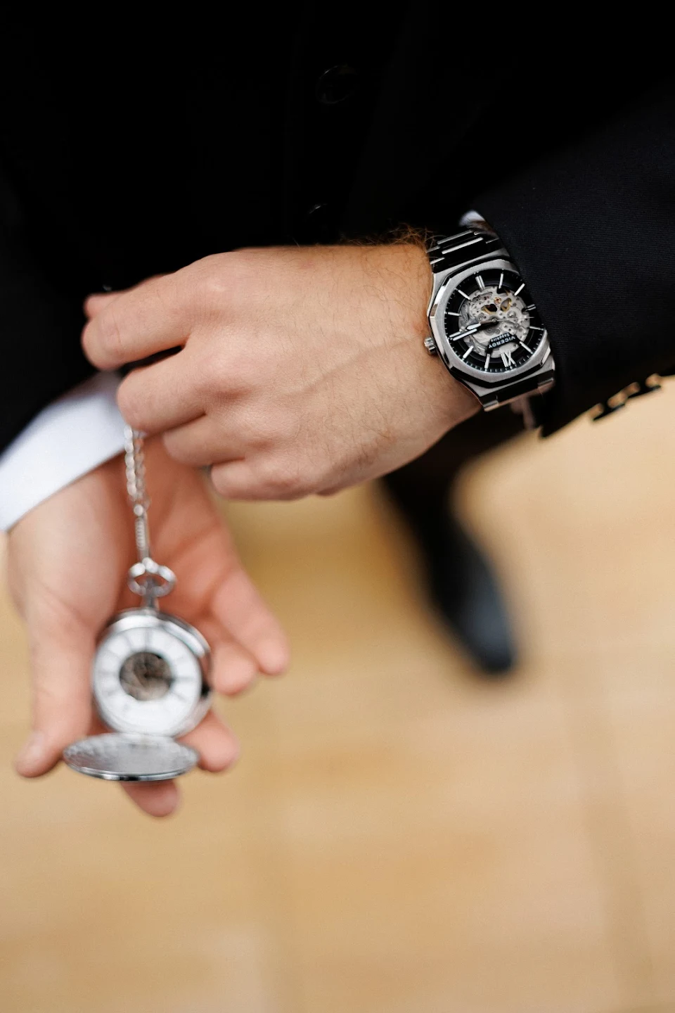 Close-up of a man’s hand wearing a skeleton wristwatch and holding an open silver pocket watch on a chain.