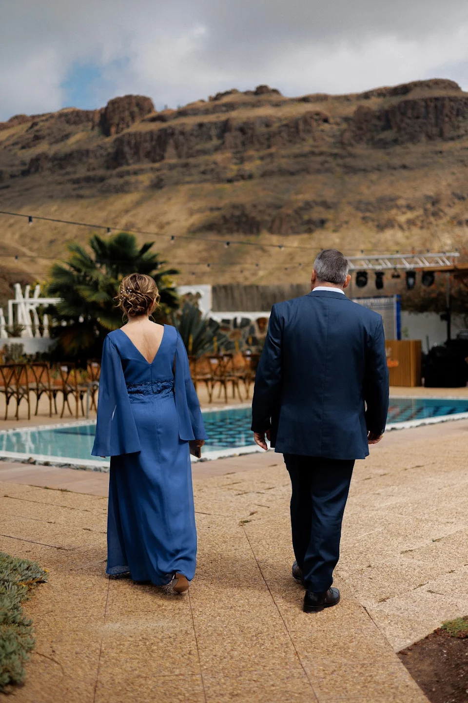A woman in a long blue dress and a man in a dark blue suit walking beside a pool with mountains in the background.