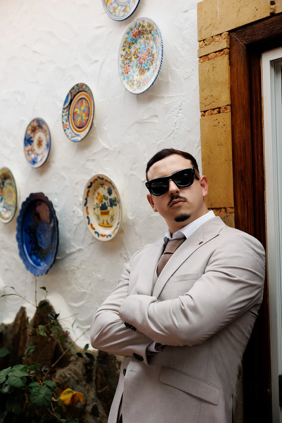 Man in a beige suit and tie wearing black sunglasses standing with crossed arms against a textured white wall decorated with colorful ceramic plates.