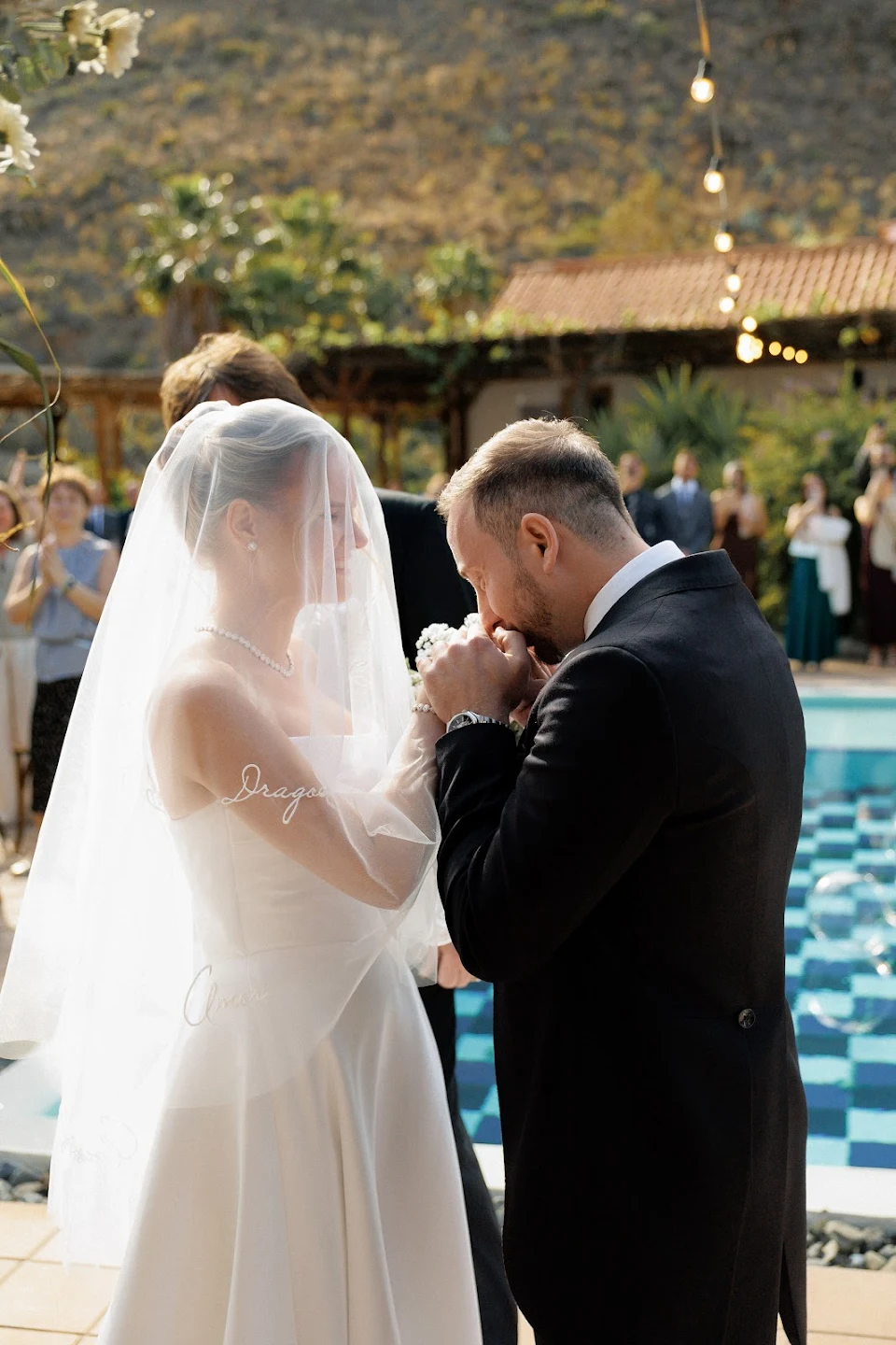Bride in white gown and veil holding hands as groom kisses her hand during outdoor wedding ceremony with guests in background.