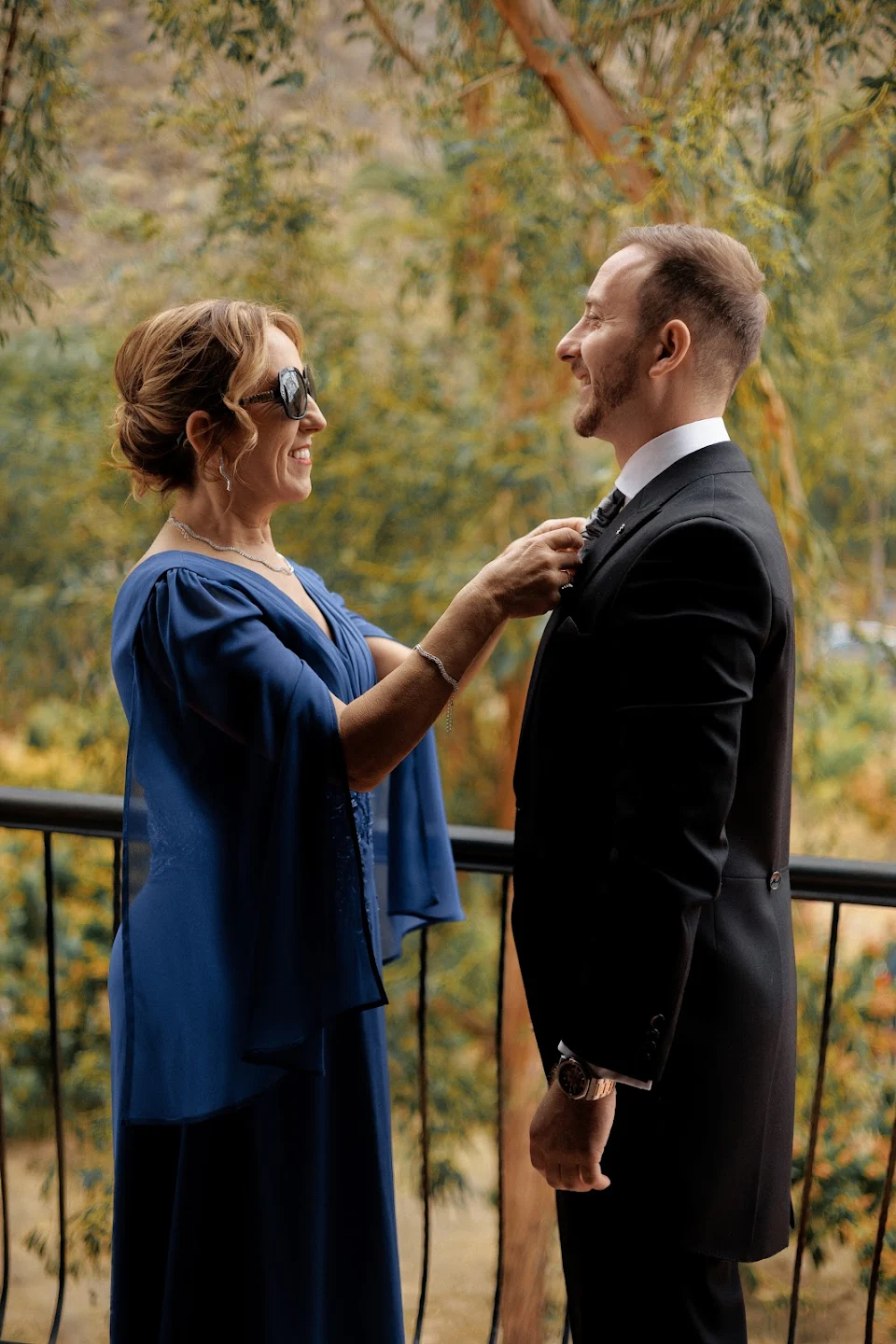 Woman in a blue dress adjusting a man's tie outdoors on a balcony with trees in the background.