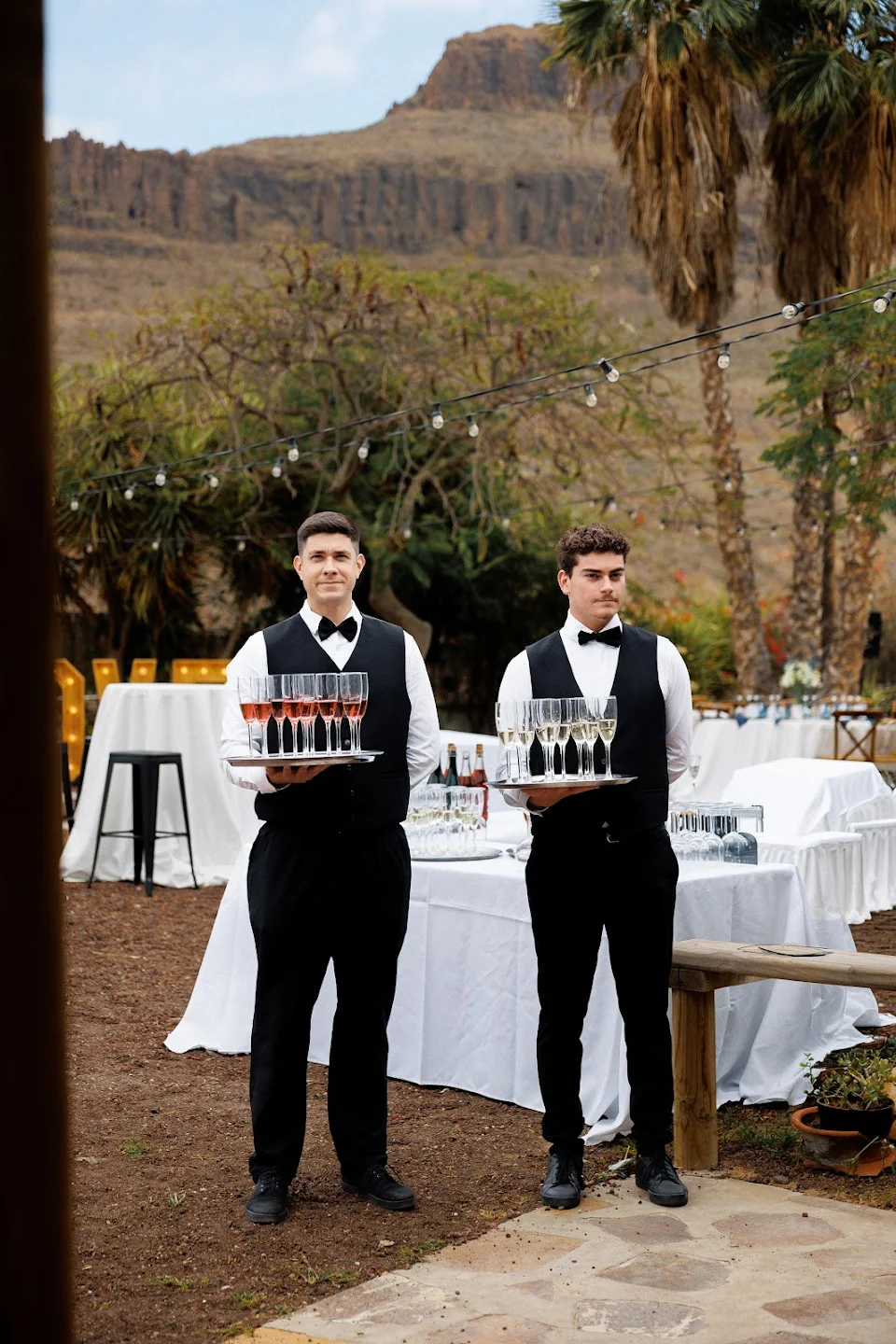 Two waiters in black vests and bow ties holding trays of champagne glasses at an outdoor event with tables and string lights.