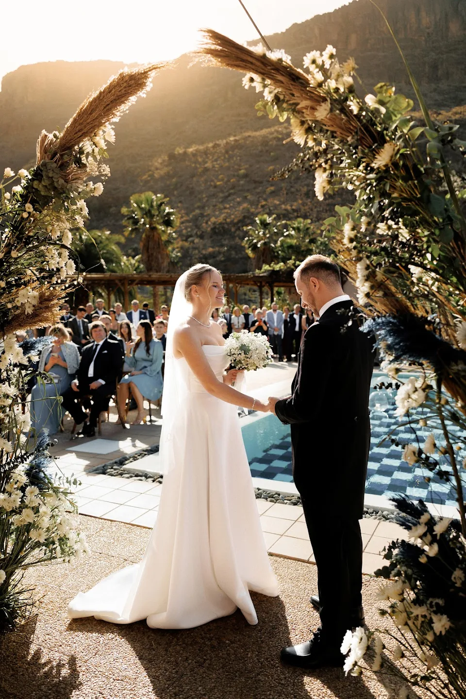 Bride and groom holding hands and facing each other under a floral arch at an outdoor wedding ceremony with guests seated in the background.