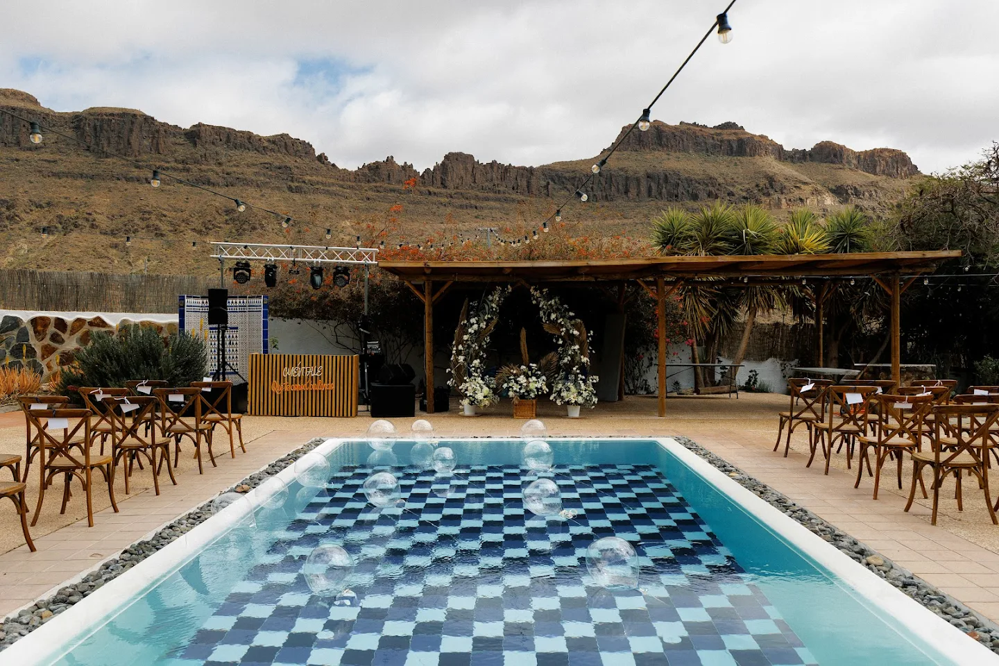 Outdoor event setup with a rectangular pool featuring a blue and black checkerboard pattern, transparent balloons floating above, wooden chairs arranged on either side, and a floral arch under a wooden pergola against a mountainous backdrop.