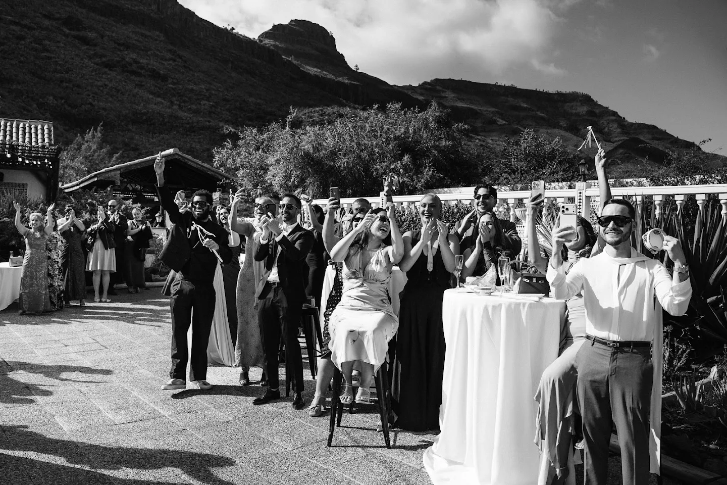 Group of formally dressed people outdoors clapping, cheering, taking photos, and celebrating against a mountainous background.
