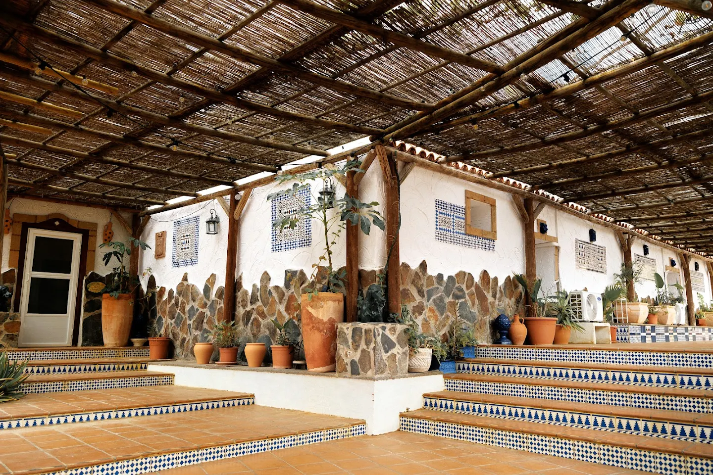 Covered outdoor terrace with stone and white walls adorned with blue tile patterns, terracotta pots with plants, and tiled steps.