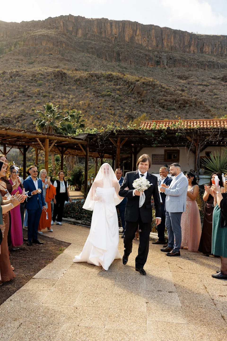 Bride in white dress and veil walking arm-in-arm with a man in a dark suit holding a bouquet, guests clapping on either side outdoors with mountainous background.