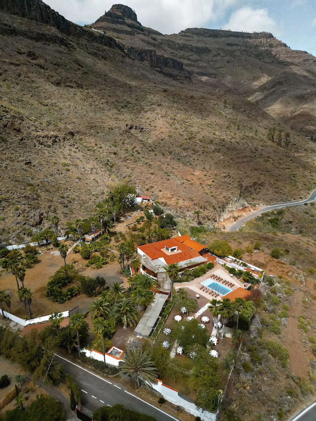 Aerial view of a hillside villa with orange roofs, surrounded by palm trees, gardens, and an outdoor swimming pool.