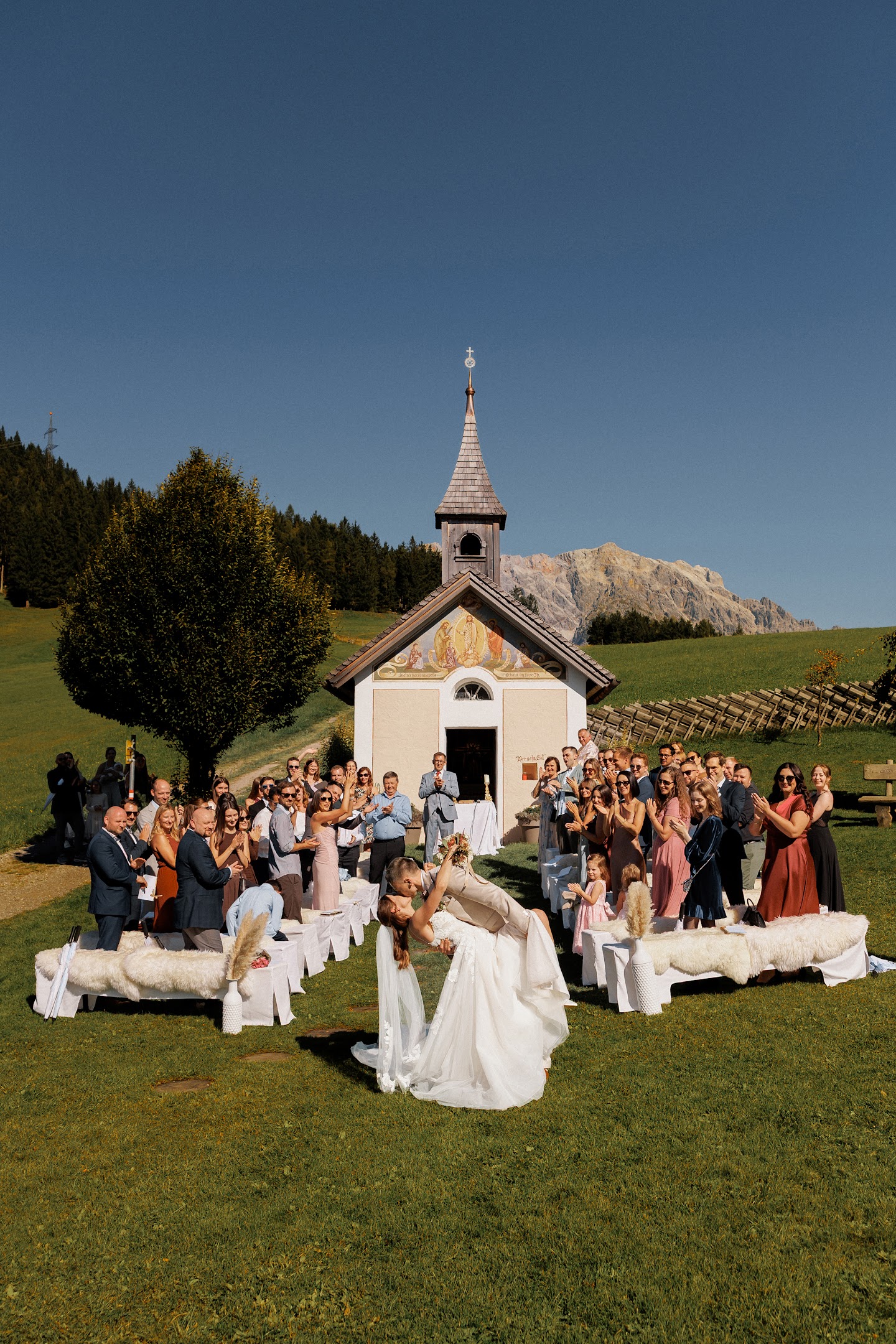Bride and groom share a kiss in front of a small chapel outdoors surrounded by guests applauding under a clear blue sky.