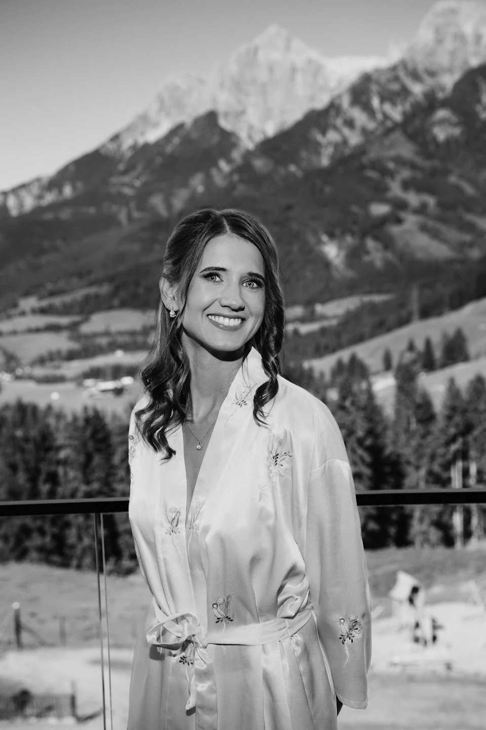 Smiling woman with curled hair wearing a floral satin robe standing outdoors with mountains and trees in the background.