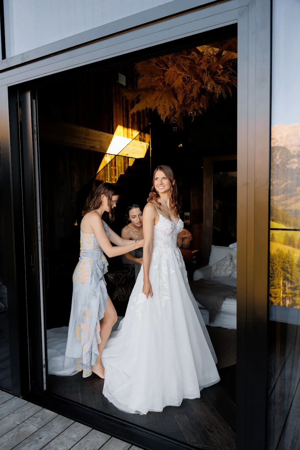 Bride in a white wedding dress smiling as two bridesmaids adjust the back of her gown inside a modern room with large glass doors.