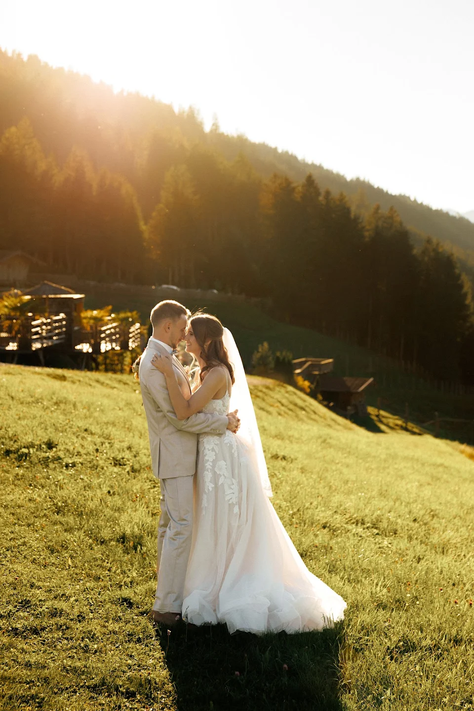 Bride and groom embracing on a sunlit grassy hillside with forested mountains in the background.