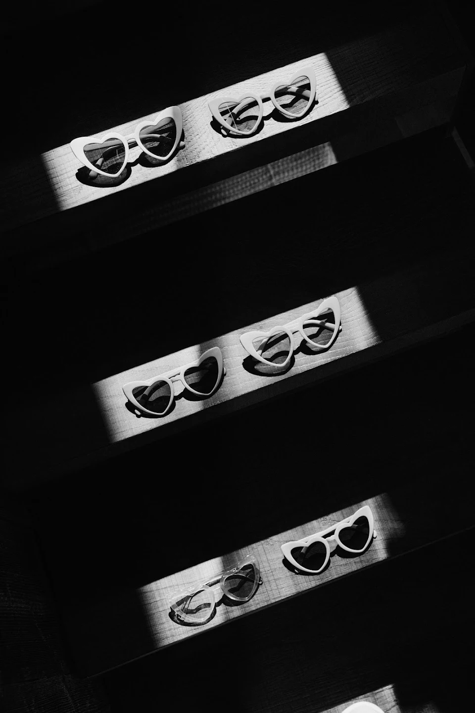Six pairs of heart-shaped sunglasses placed on wooden steps under strips of sunlight in a black and white photo.