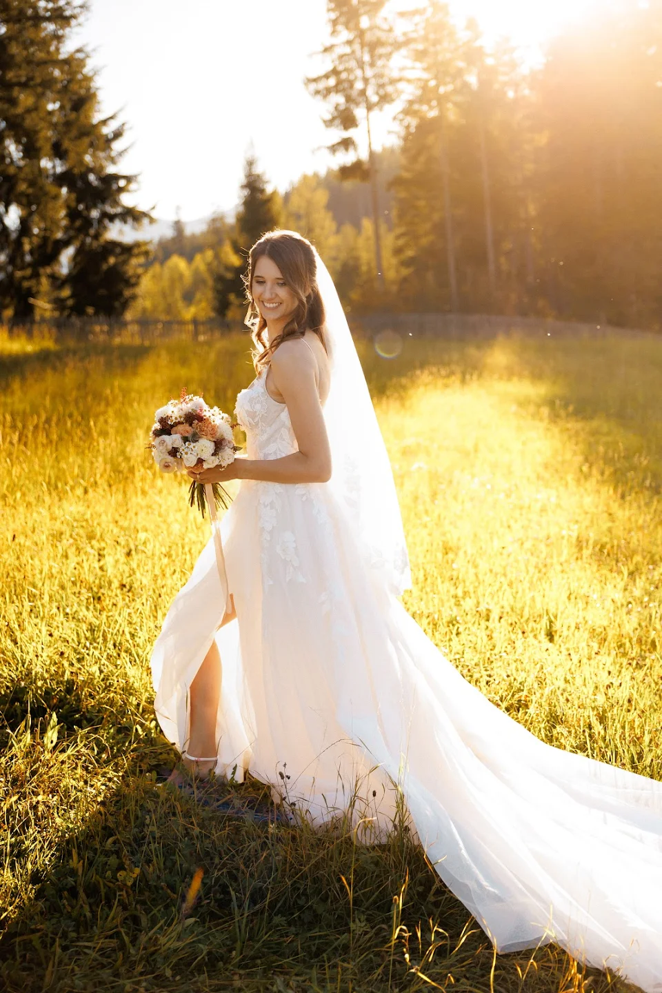 Bride in a white wedding gown holding a bouquet, standing in a sunlit grassy field with trees in the background.